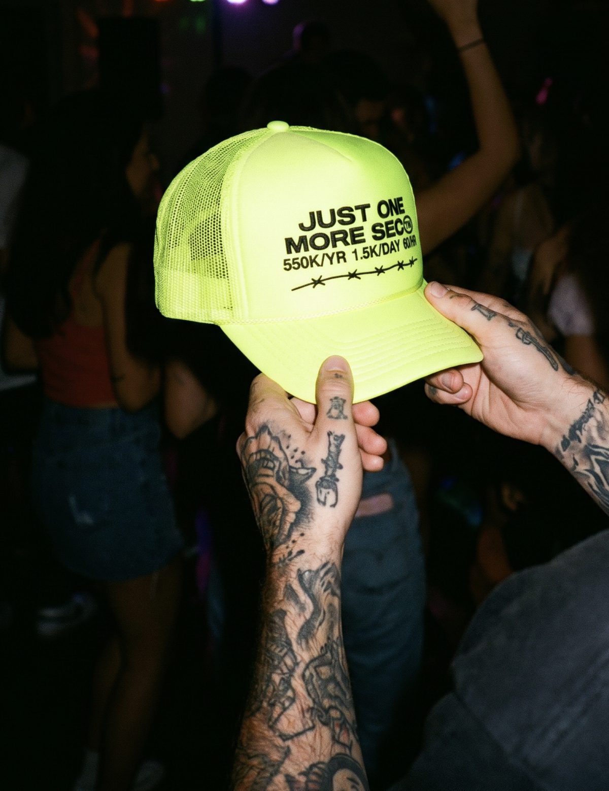 A hand holding a neon SIXTY-ONE Just One More Sec trucker hat in a crowd